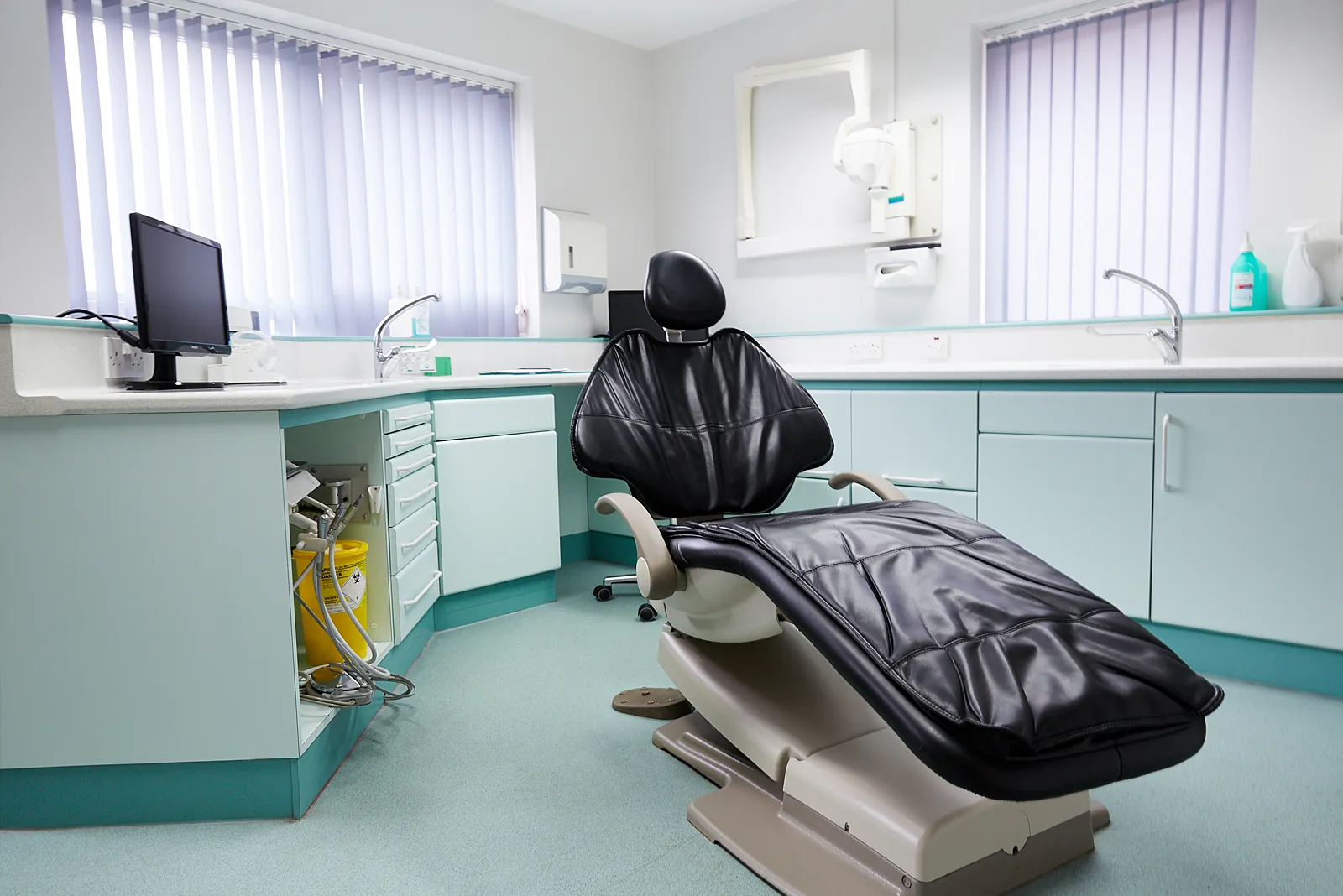 A bright treatment room with a black dental chair and sea-green cabinets